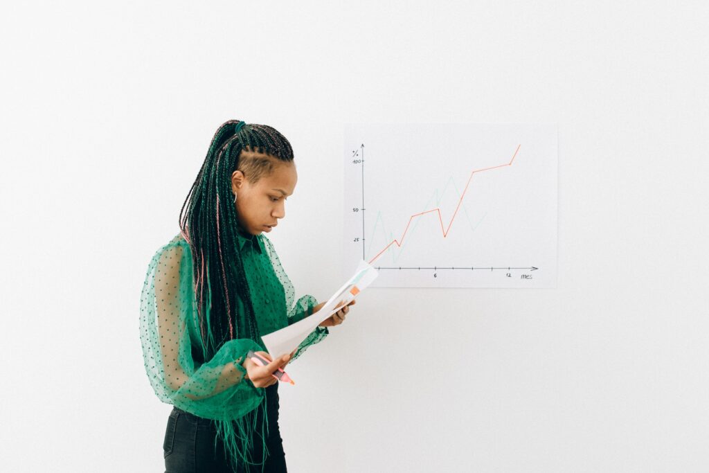 A person with braids and a green blouse studying a sheet of paper. Behind them is a graph showing an upward-trending red line.